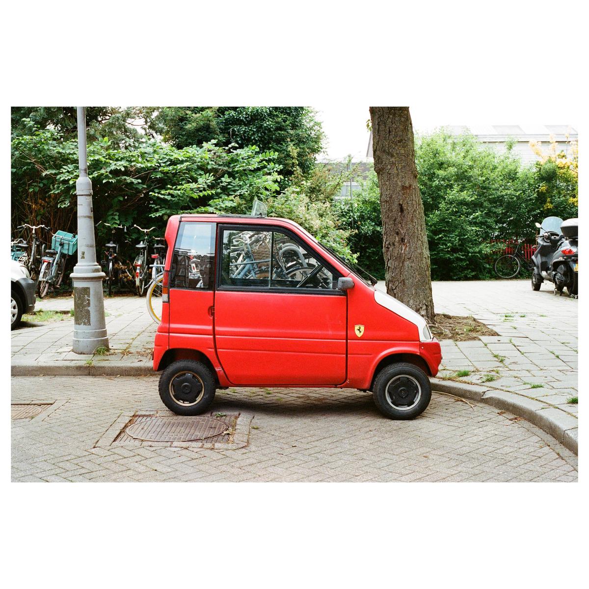 A small red car is parked on a paved street next to a tree and a lamppost.
