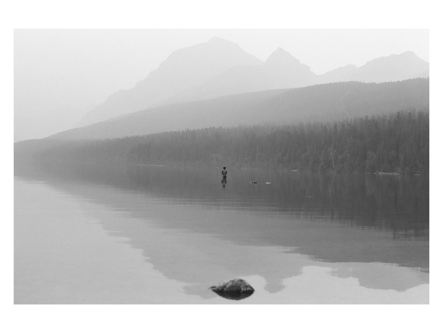 A person stands in a hazy lake with mountains and a forest in the background.
