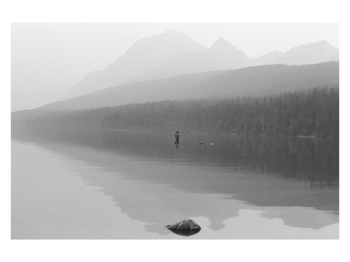 A person stands in a hazy lake with mountains and a forest in the background.