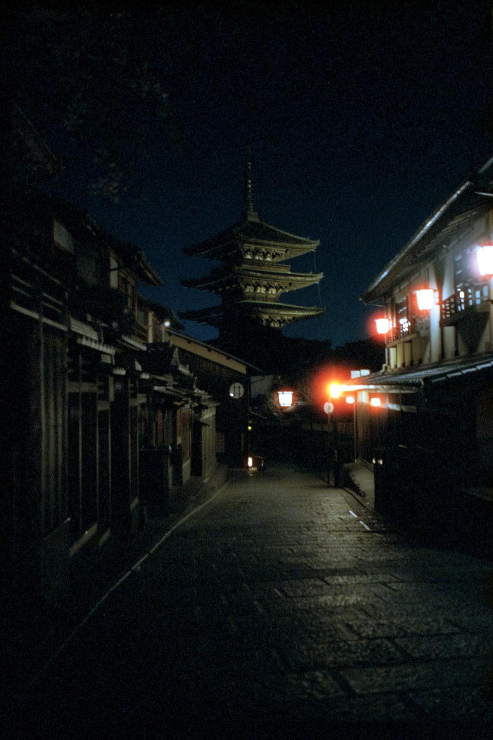 A narrow street with traditional buildings and a pagoda in the distance is illuminated by lanterns at night.