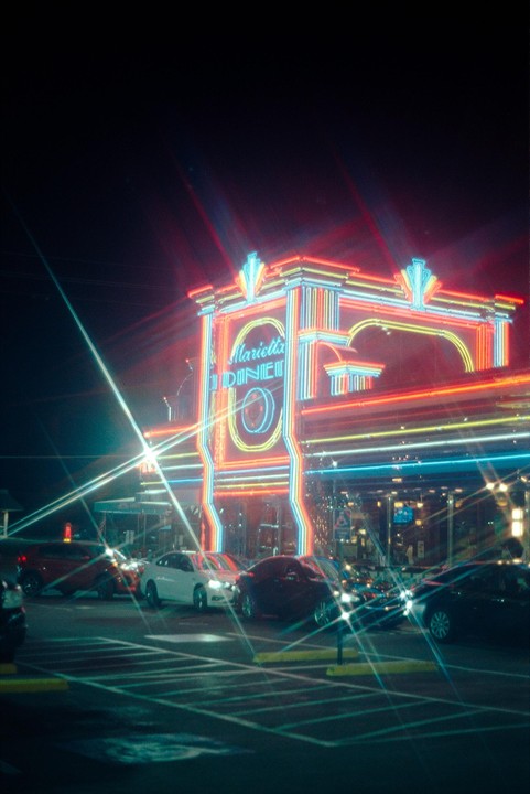 A diner with a brightly lit neon sign stands at night with cars parked in front.