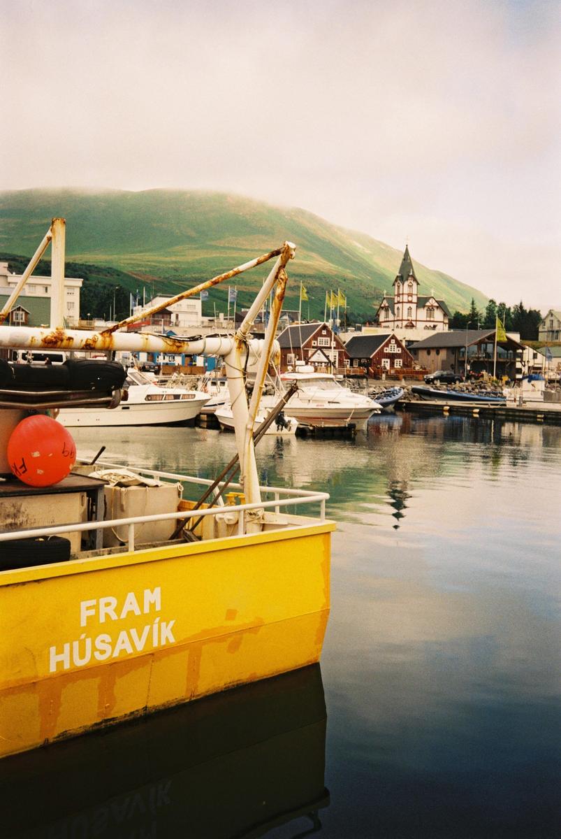 A yellow boat is docked in a harbor with buildings and a green mountain in the background.