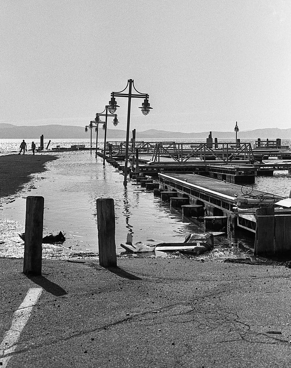 Two people walk along a flooded shore next to a row of empty boat docks and street lamps.