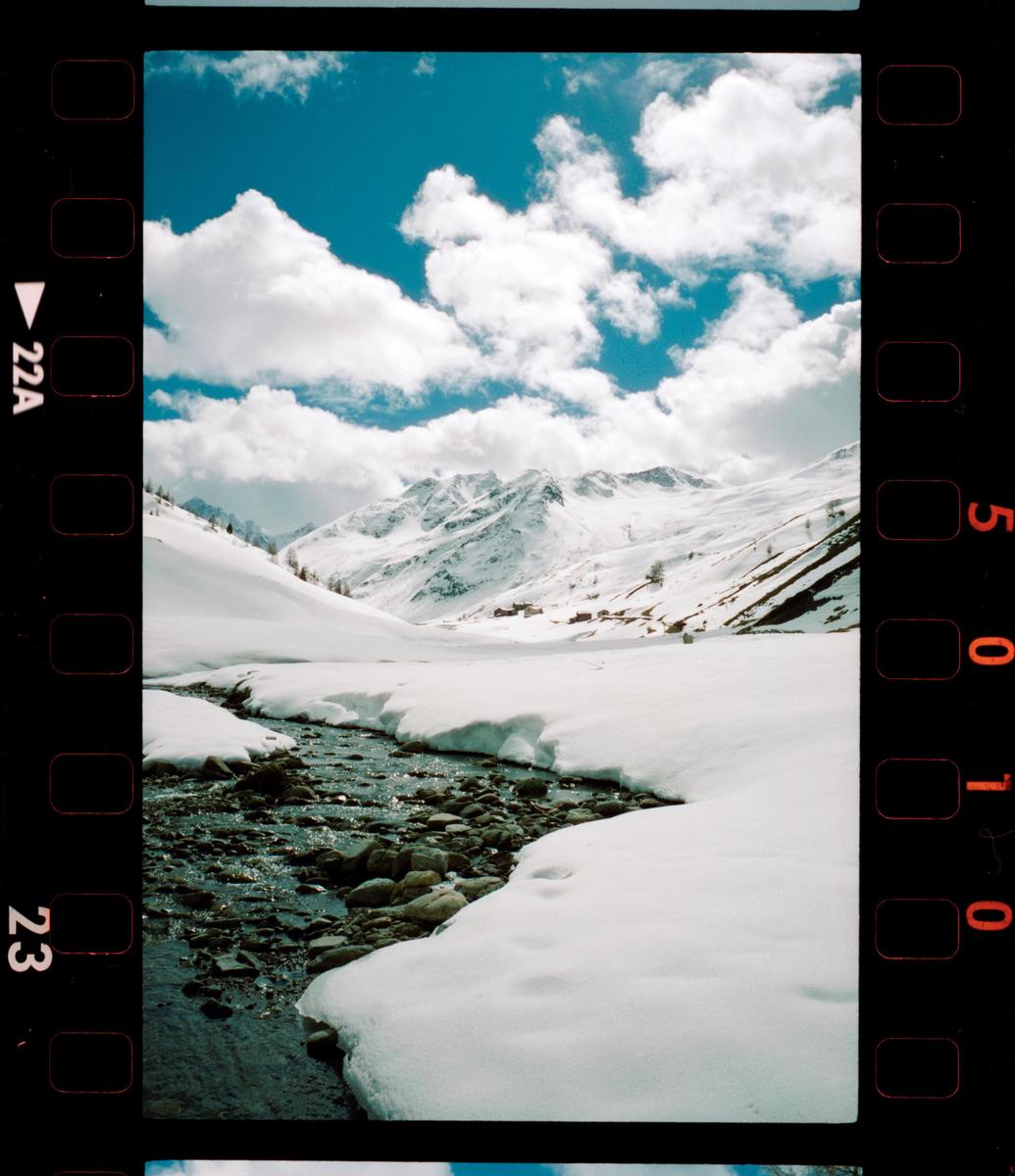 A stream flows through a snow-covered valley with mountains in the background under a blue sky with clouds.