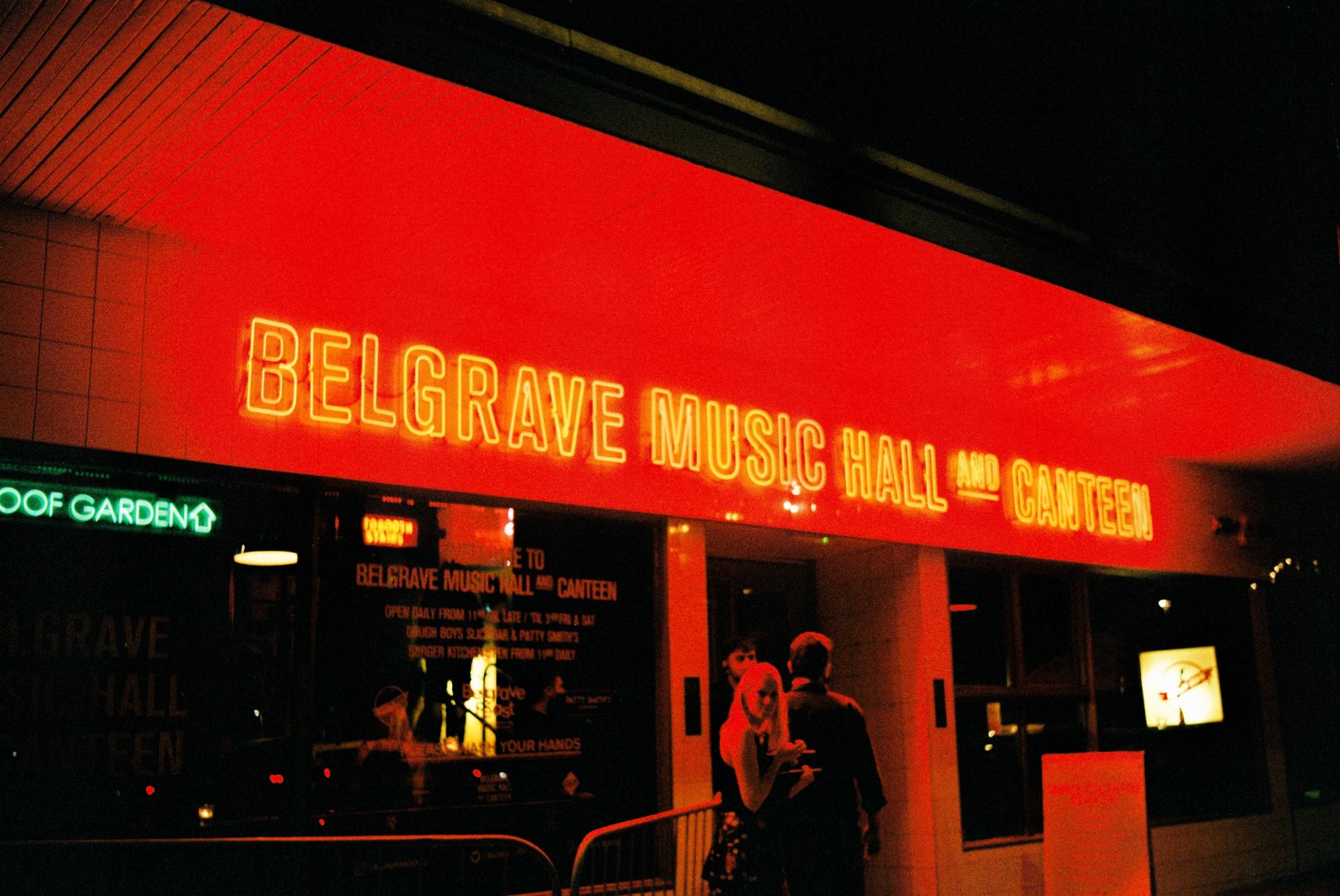A neon sign for Belgrave Music Hall and Canteen glows red above a street at night where a couple stands.