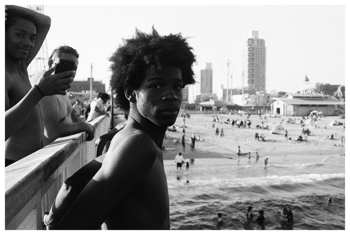 A young man with an afro looks at the camera from a pier overlooking a crowded beach with buildings in the background.