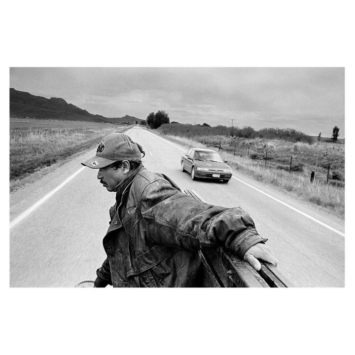 A man in a cap and jacket holds onto the back of a moving vehicle on a rural road with another car approaching in the distance.