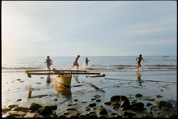 Four children run and play in the shallow water of a rocky beach next to a small boat.