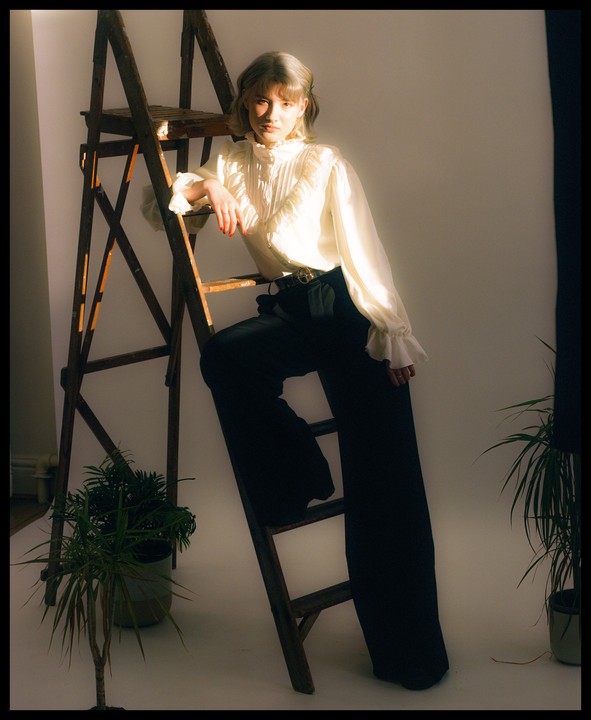 A woman in a white ruffled blouse and black pants leans on a wooden ladder next to a potted plant.