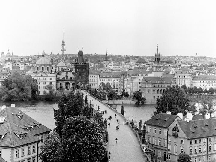 An elevated view shows a bridge with people walking across it, leading to a city with many buildings and a river.