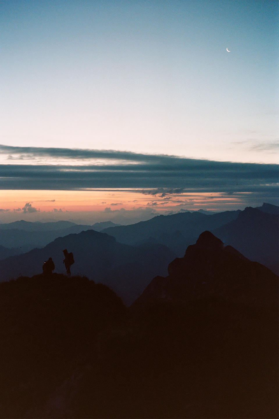 Two hikers stand on a mountain ridge at dawn with a crescent moon visible in the sky.