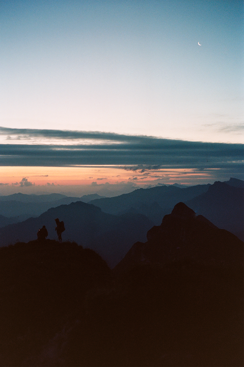 Two hikers stand on a mountain ridge at dawn with a crescent moon visible in the sky.