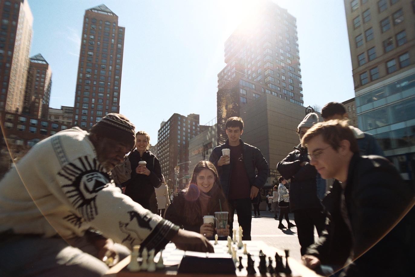 A man plays chess with a young woman on a city street with tall buildings in the background.