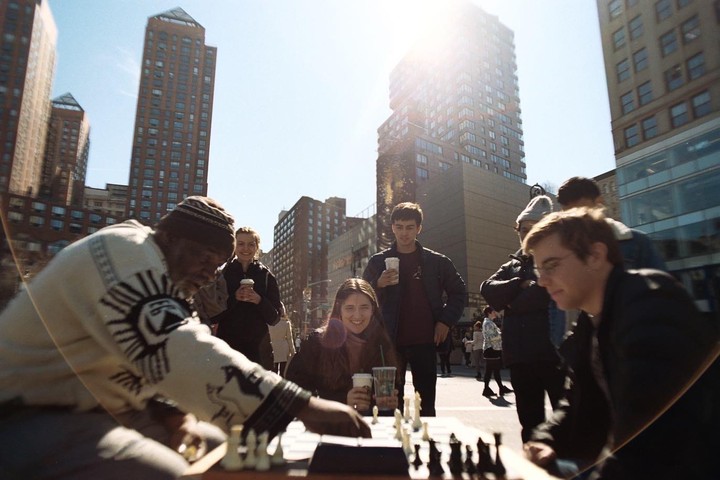 A man plays chess with a young woman on a city street with tall buildings in the background.