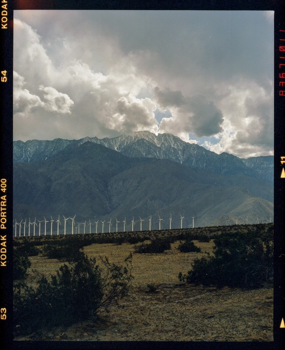 A field of wind turbines stands before a mountain range with snow-capped peaks under a cloudy sky.
