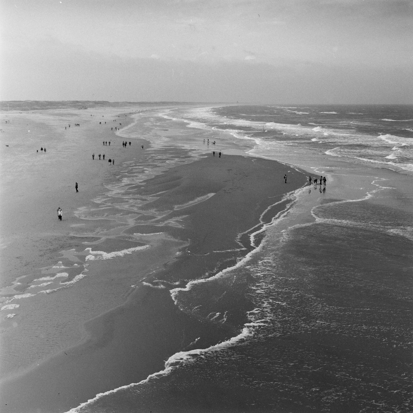 A high-angle view shows a long beach with many people walking along the shore and waves breaking in the ocean.