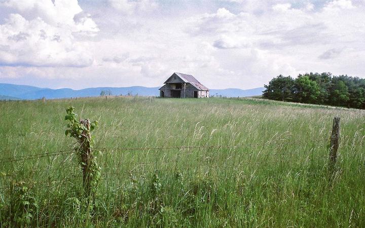 An old barn stands in a field of tall grass with mountains in the background under a cloudy sky.