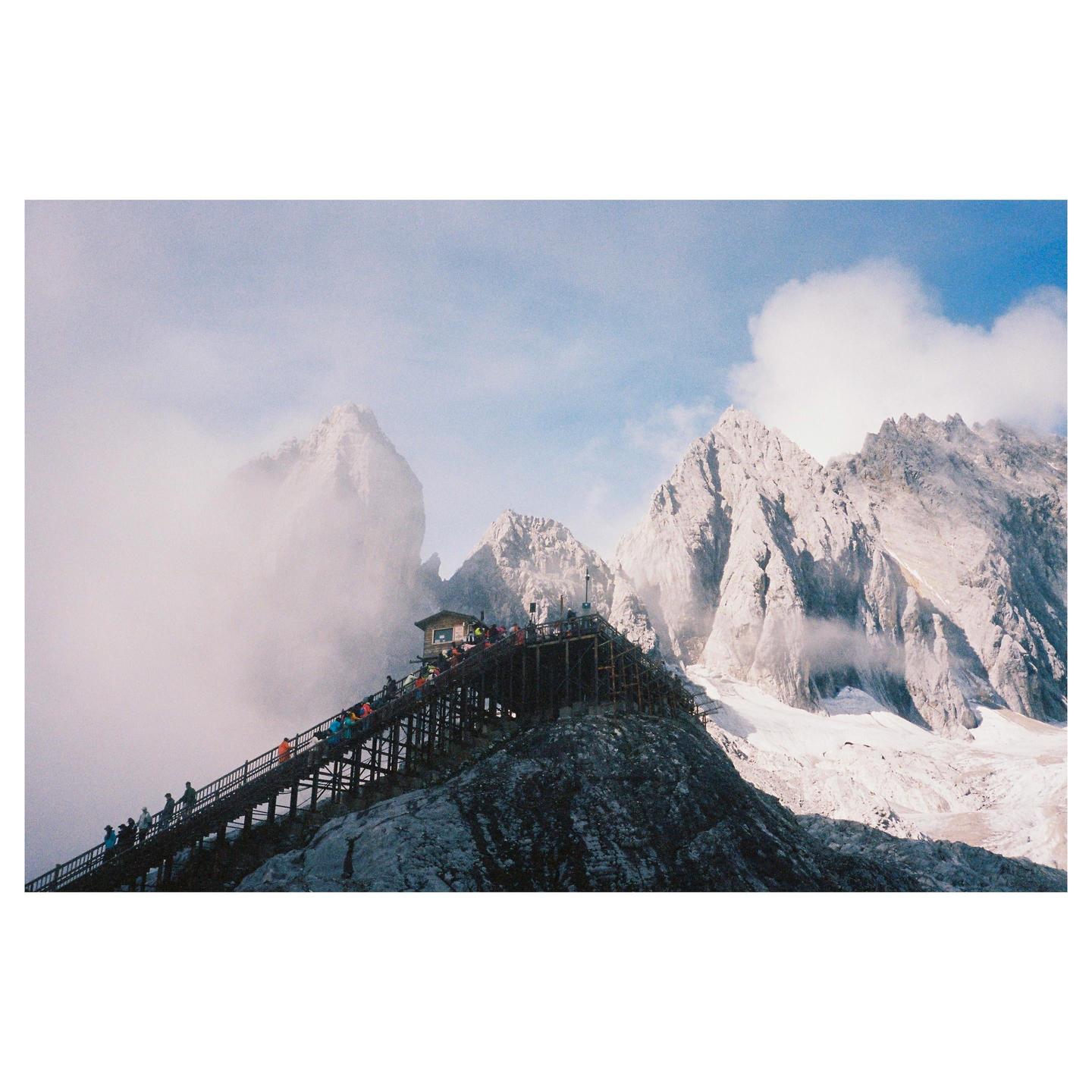 A long wooden staircase with many people leads up a mountain towards a small building, with snow-capped peaks and clouds in the background.