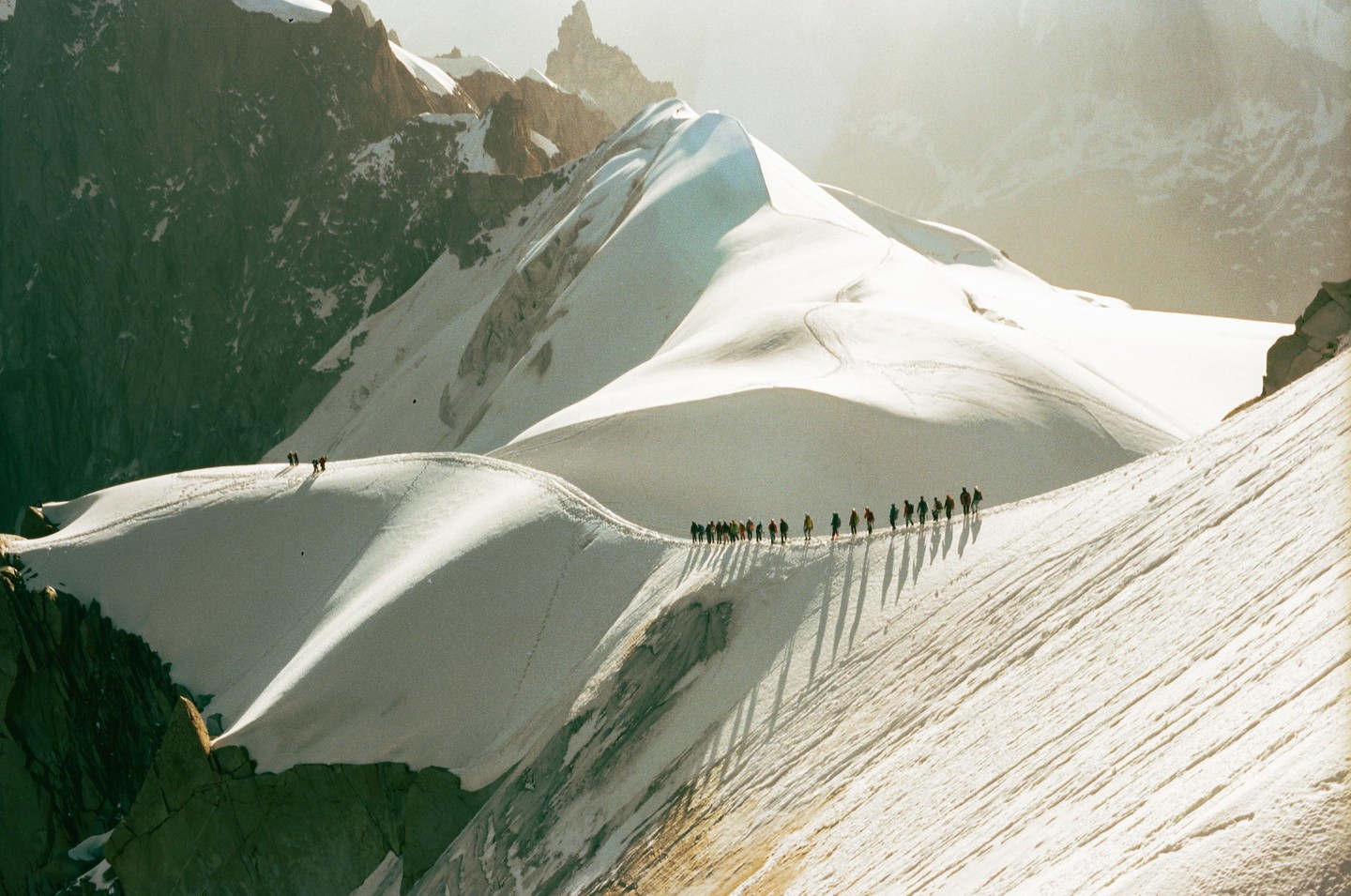A line of alpinists with long shadows walks across a snow-covered mountain ridge with other peaks in the background.