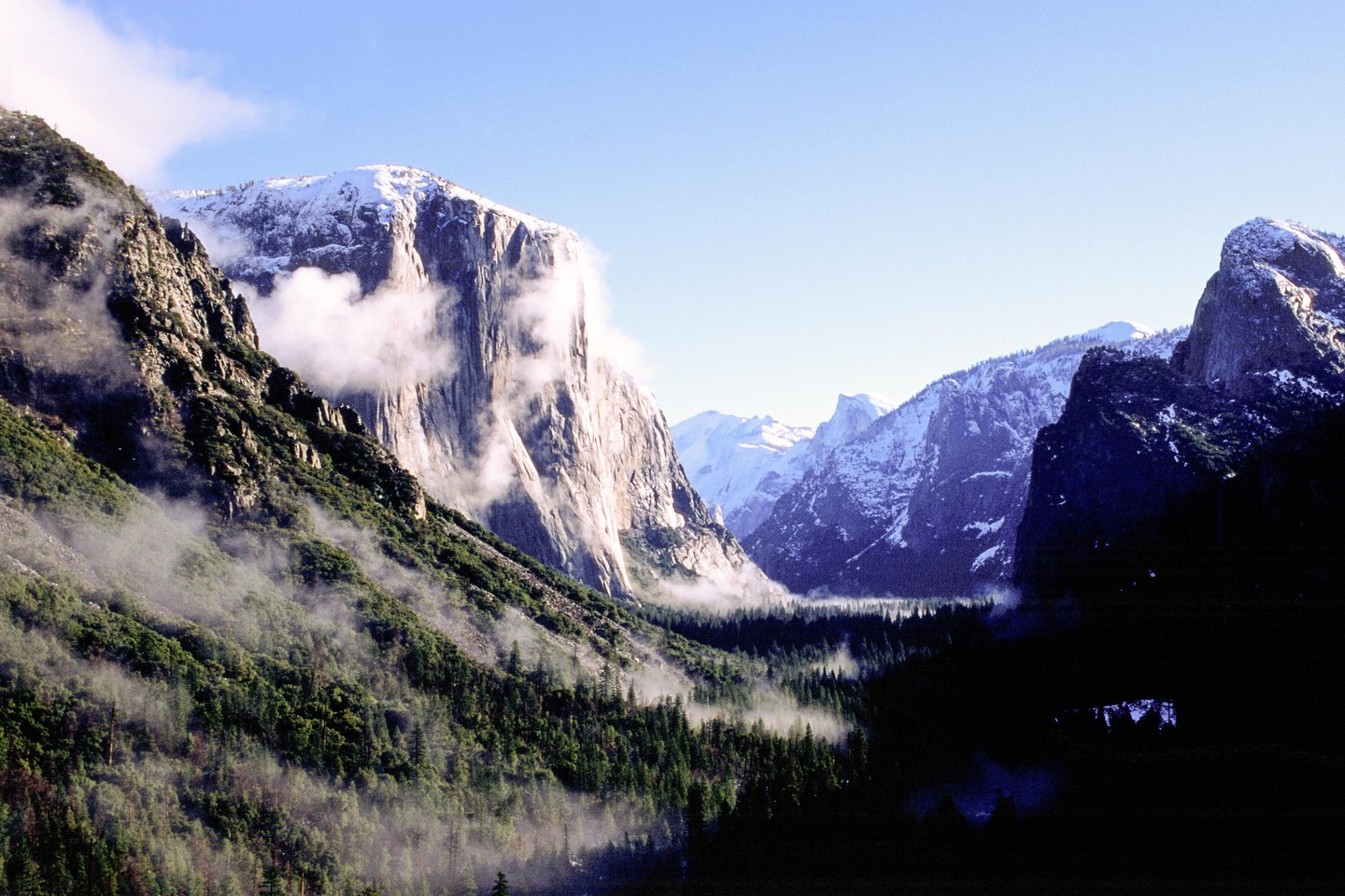 Fog clears from a valley surrounded by mountains with snow-capped peaks and evergreen trees.