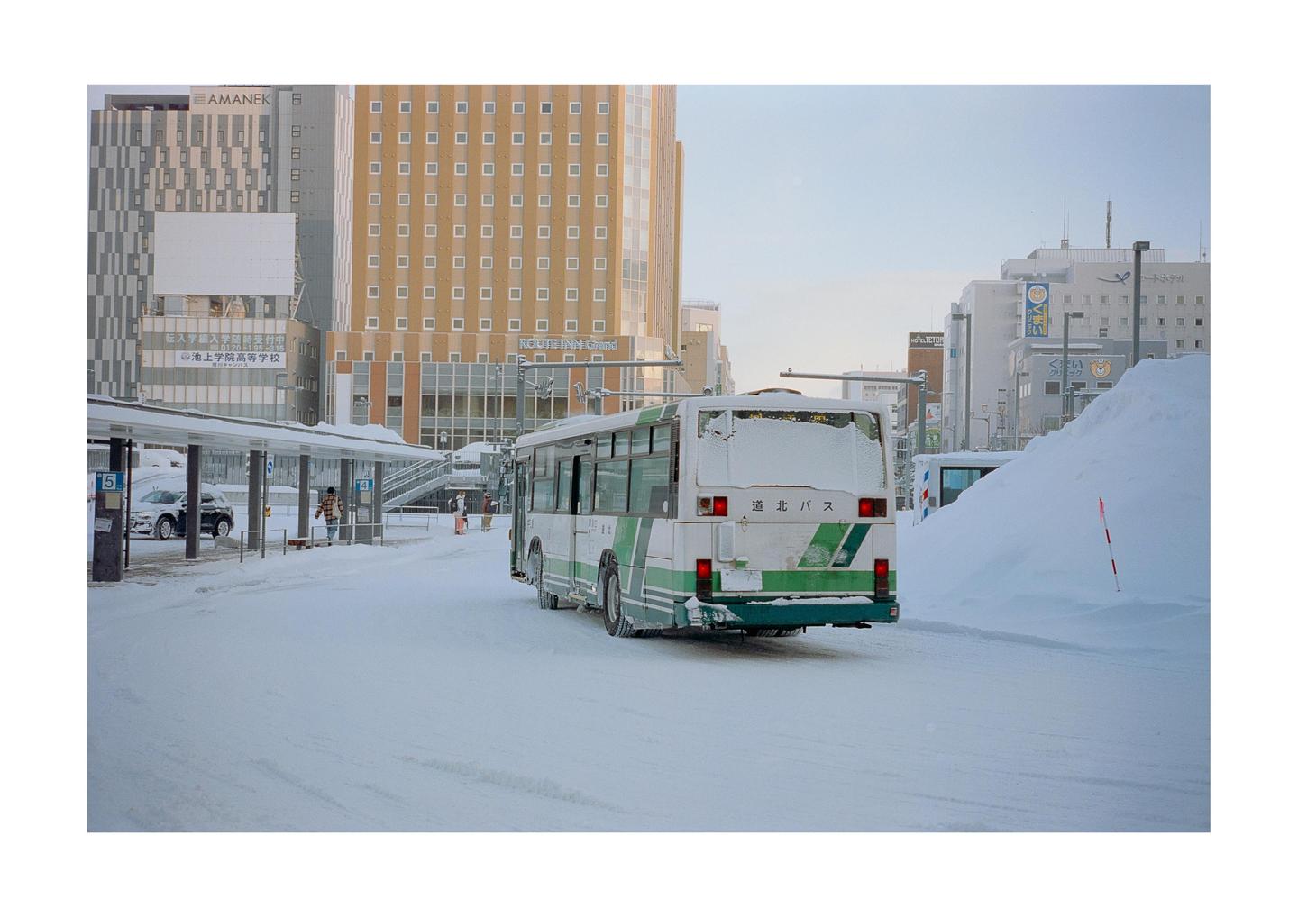 A white and green bus is parked on a snow-covered street in front of tall buildings.