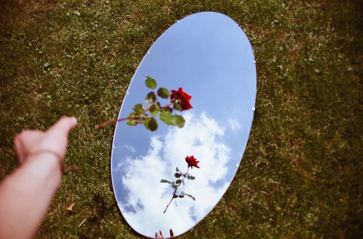 A hand points towards an oval mirror reflecting a blue sky with clouds and two red roses, all resting on green grass.
