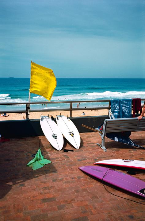 A yellow flag stands on a railing overlooking a beach with surfboards on a brick patio in the foreground.