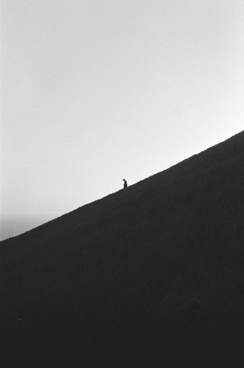 A lone figure sits on a grassy hill under a bright sky.