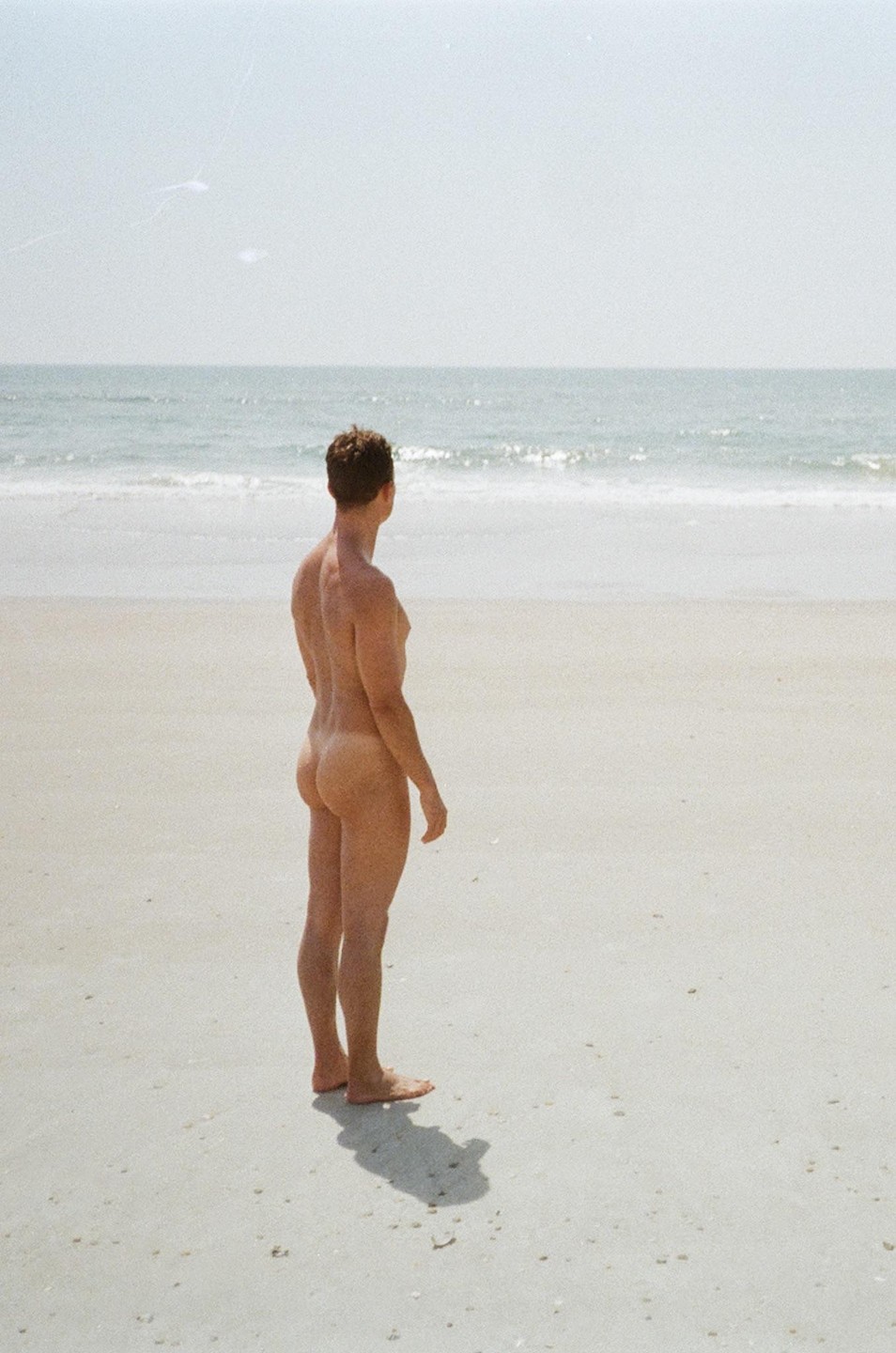 A man stands naked on a sandy beach looking out at the ocean.