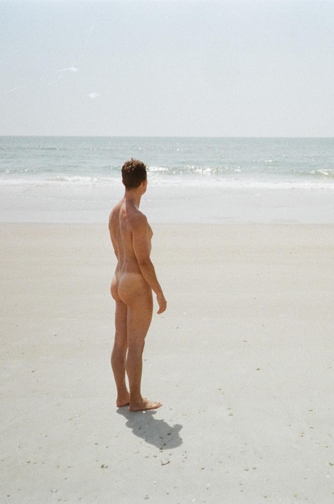 A man stands naked on a sandy beach looking out at the ocean.