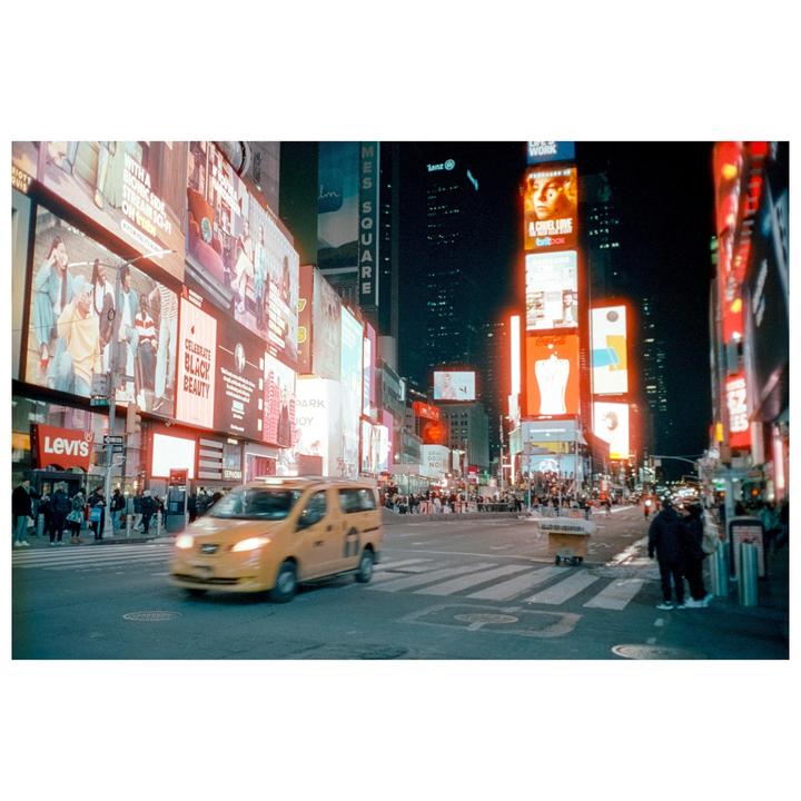A yellow taxi drives through a busy intersection at night, surrounded by towering buildings adorned with bright billboards.