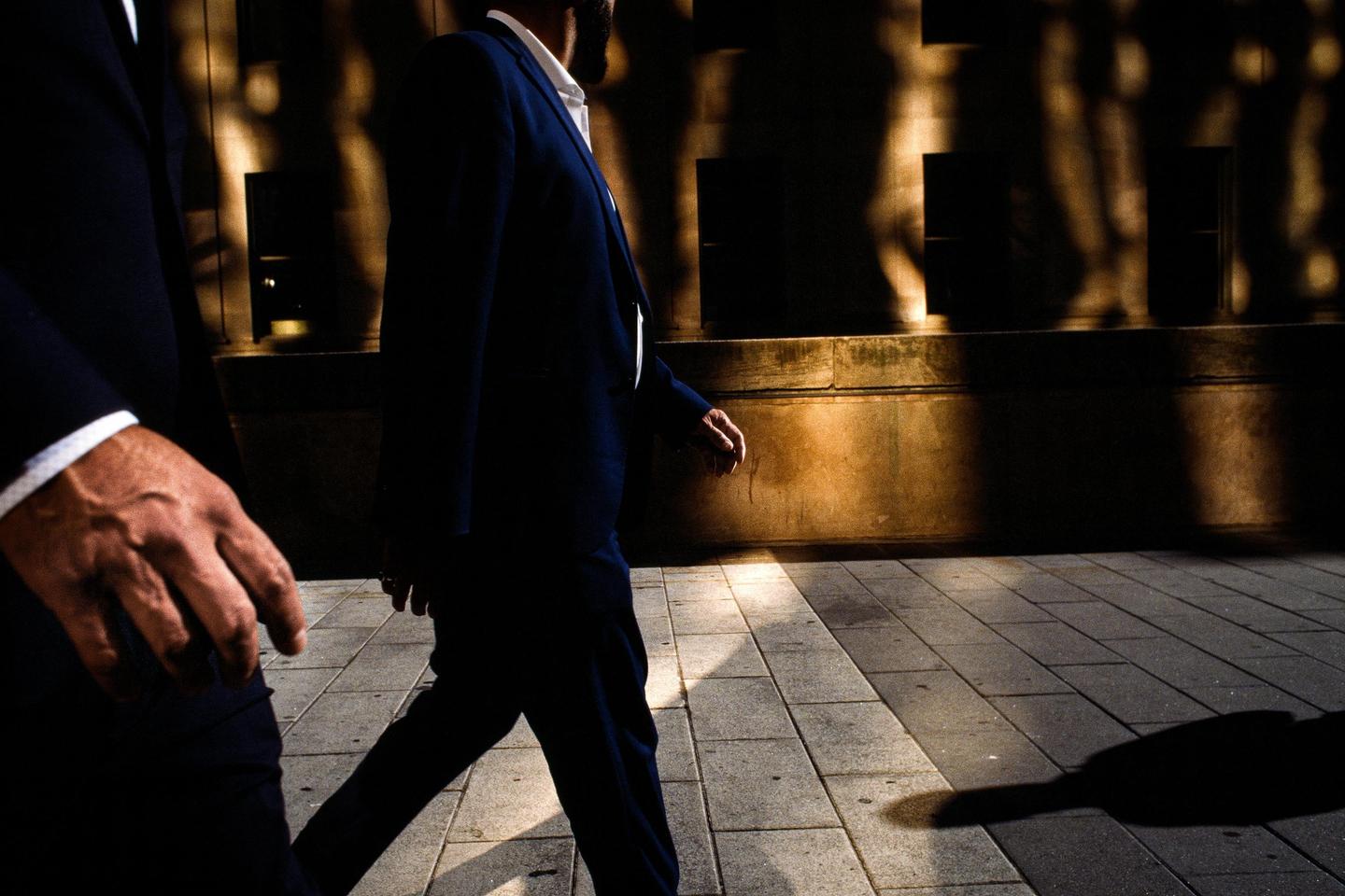 Two men in suits walk on a sidewalk with strong shadows and light patterns on a building wall.