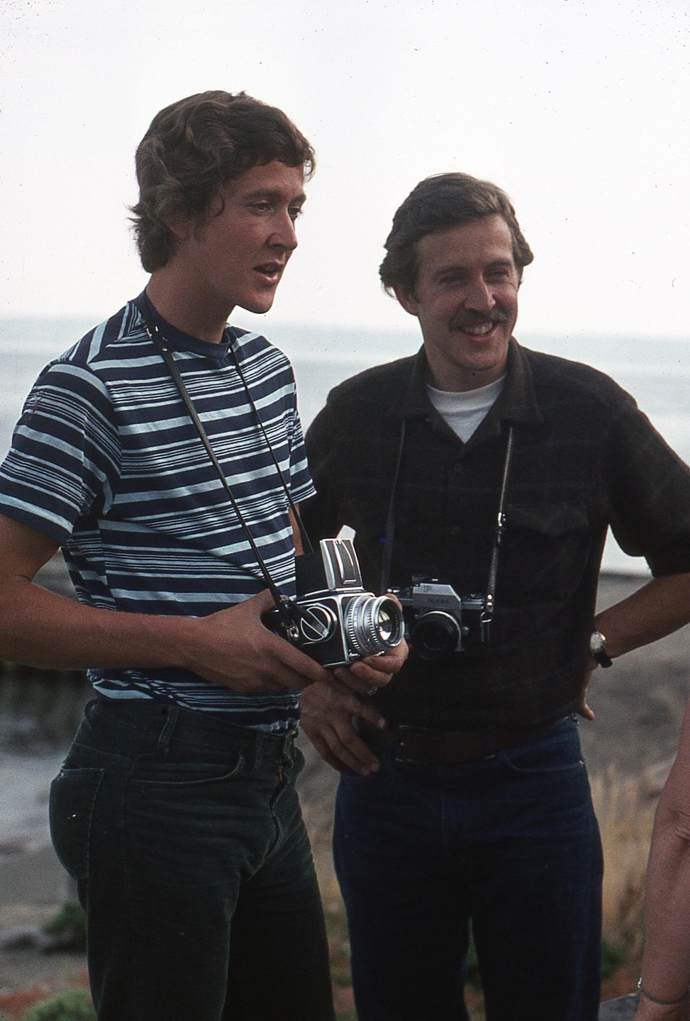 Two men stand outdoors, one holding a camera and the other smiling.
