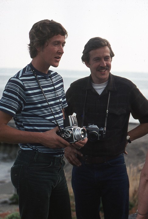 Two men stand outdoors, one holding a camera and the other smiling.