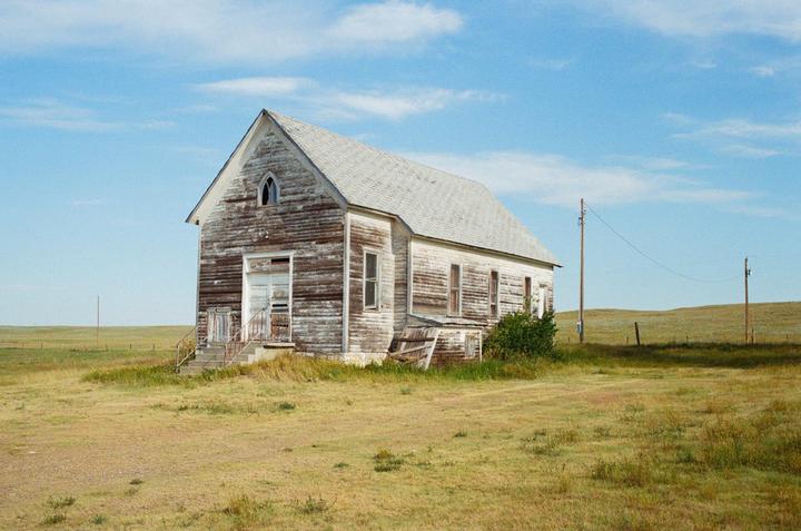 An old, weathered church stands alone in a vast, grassy prairie under a clear blue sky.