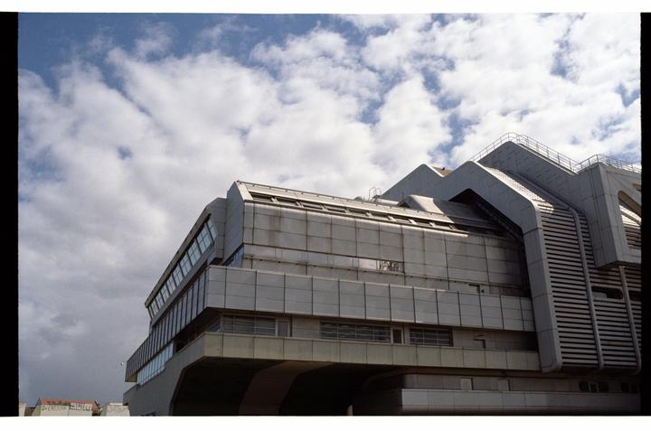 A large, angular brutalist building with many windows is shown against a cloudy sky.