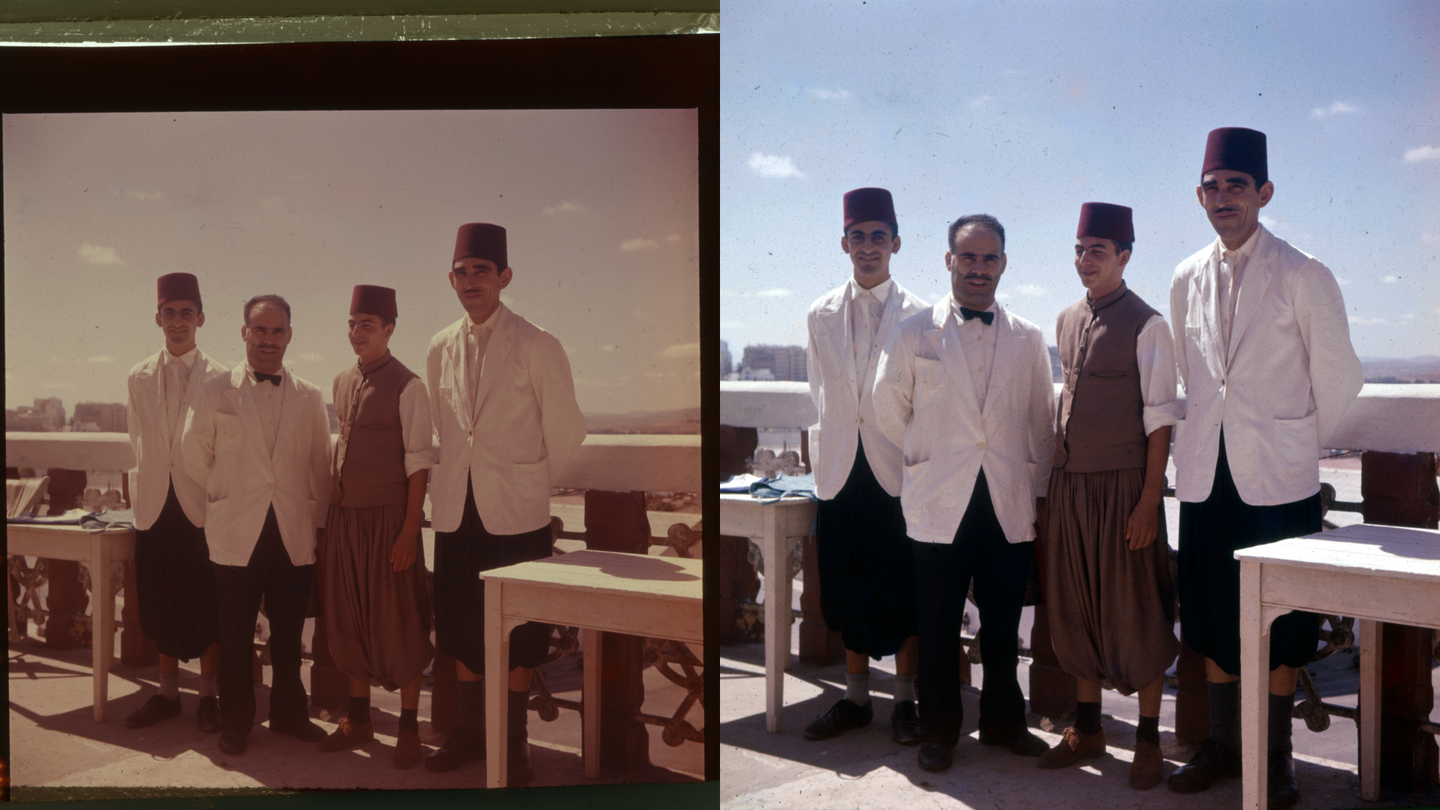 Four men in traditional attire stand on a rooftop with a city and body of water in the background.