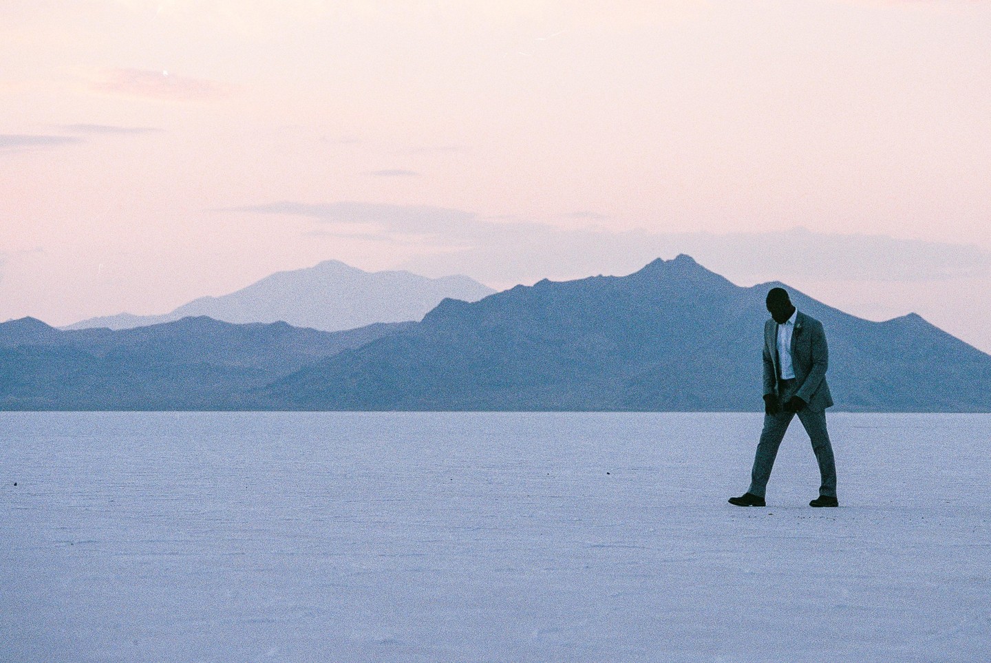 A man in a suit walks across a vast salt flat with mountains in the background under a pale sky.