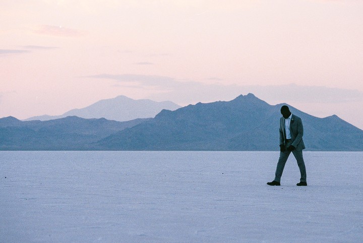A man in a suit walks across a vast salt flat with mountains in the background under a pale sky.