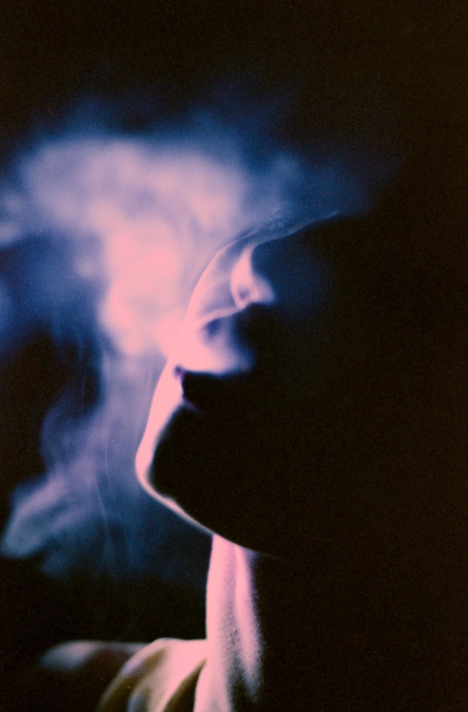 A person's profile is illuminated by pink and blue light, with smoke or vapor rising in front of their face.