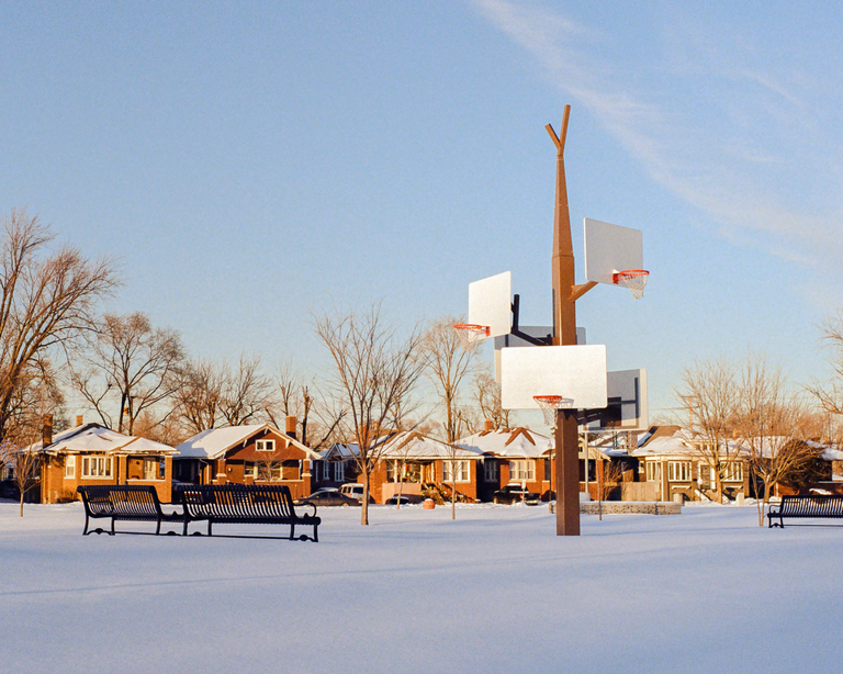 A snow-covered park with a multi-hoop basketball pole and benches is surrounded by houses and bare trees under a clear sky.