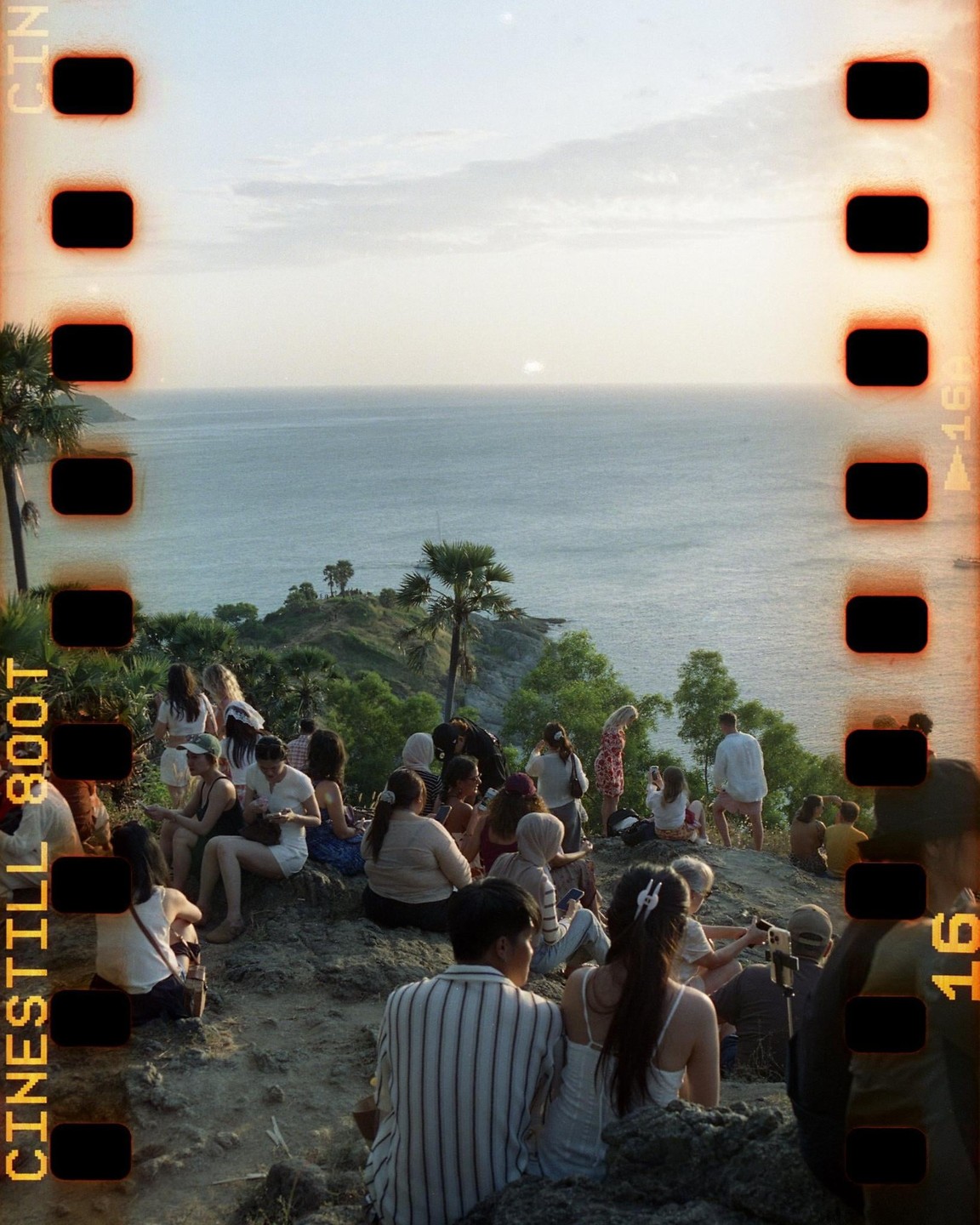 A crowd of people sits on a rocky cliff overlooking the ocean at sunset.