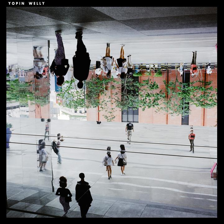 People walk on a street with reflections of buildings and trees on a glass surface.