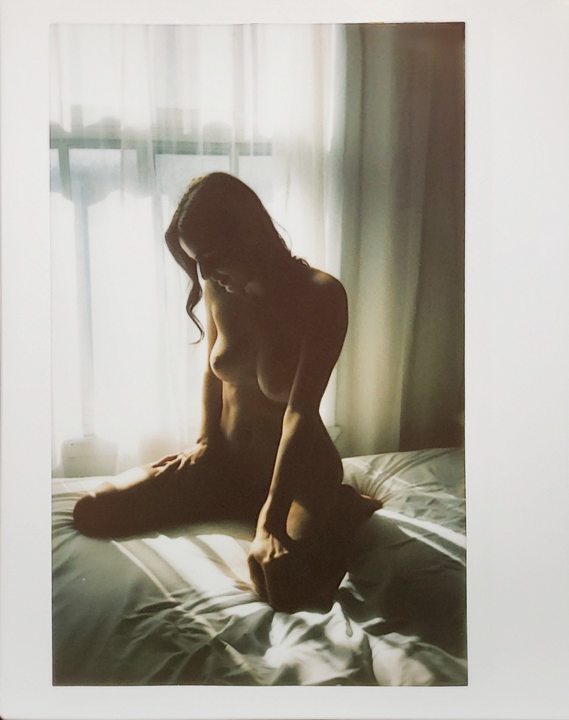 A nude woman kneels on a bed with light coming through a window behind her.