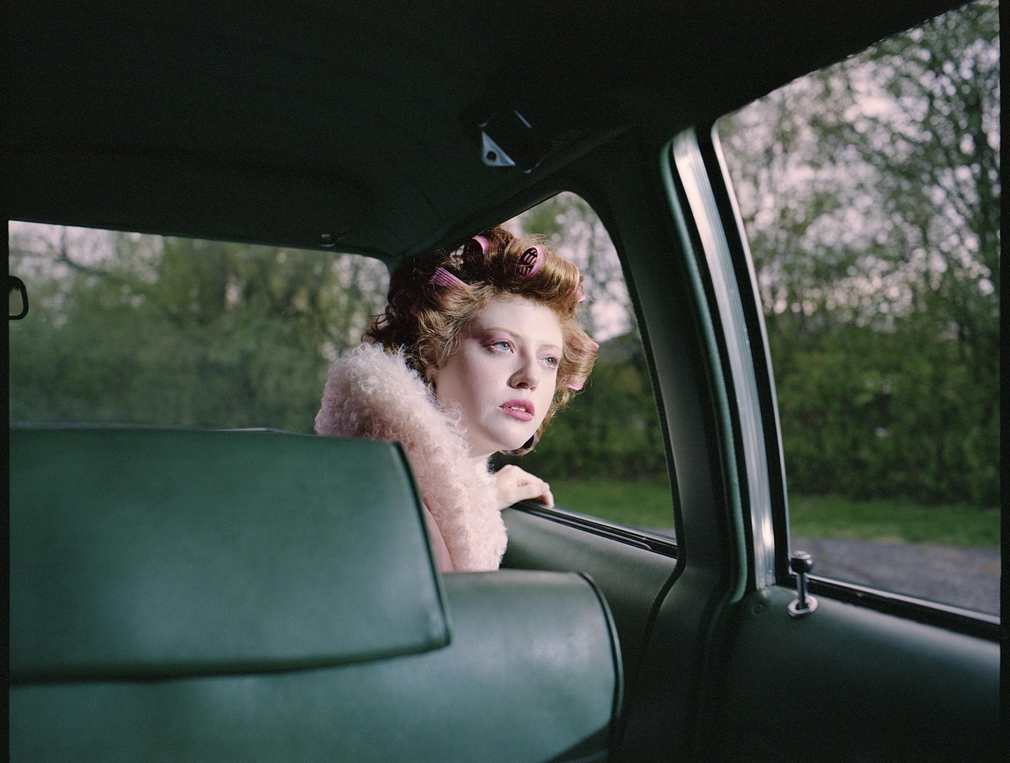 A woman with hair curlers looks out the window of a car.