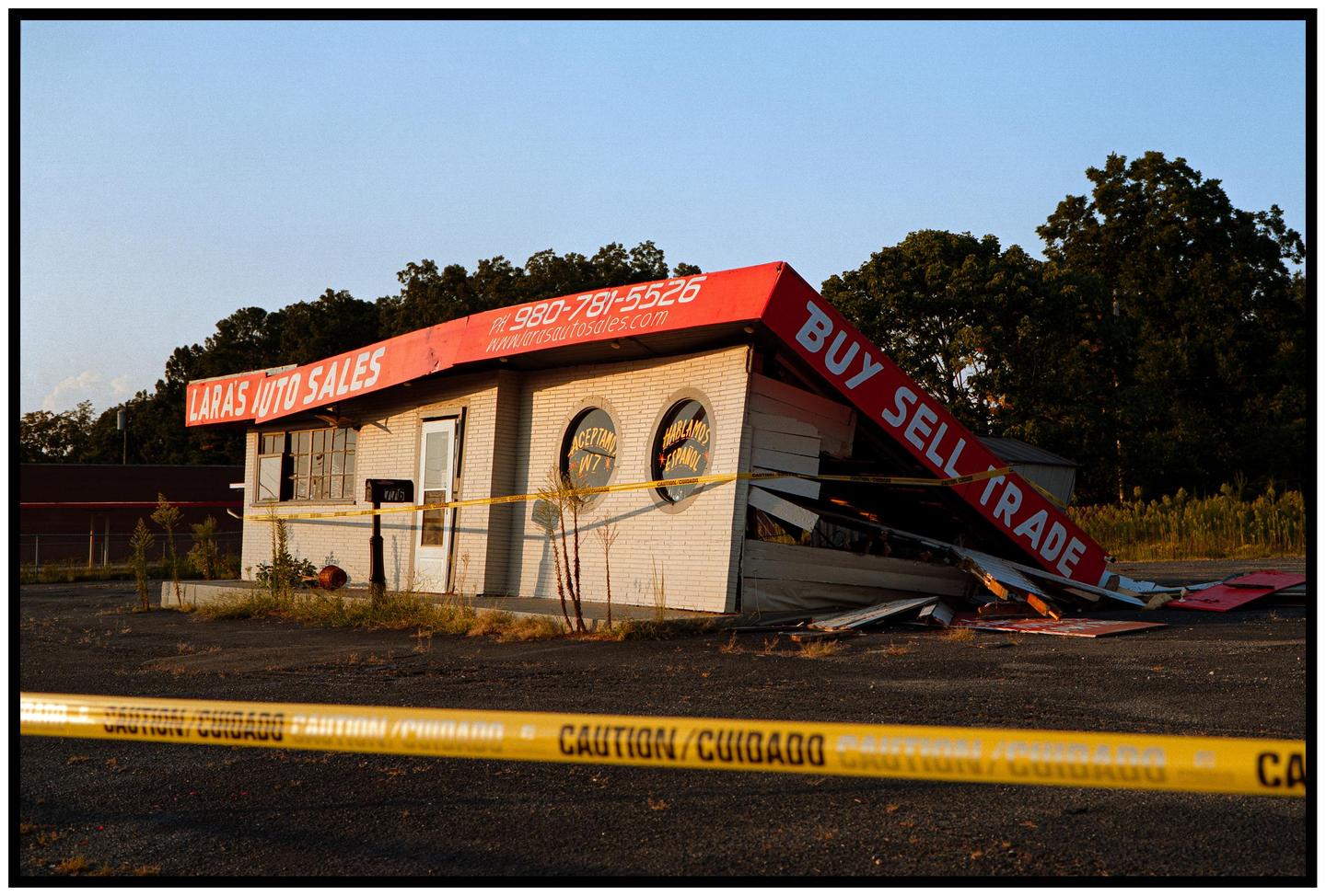 A yellow caution tape is stretched across the foreground of a damaged building with a red awning that reads "Lara's Auto Sales" and "Buy Sell Trade."