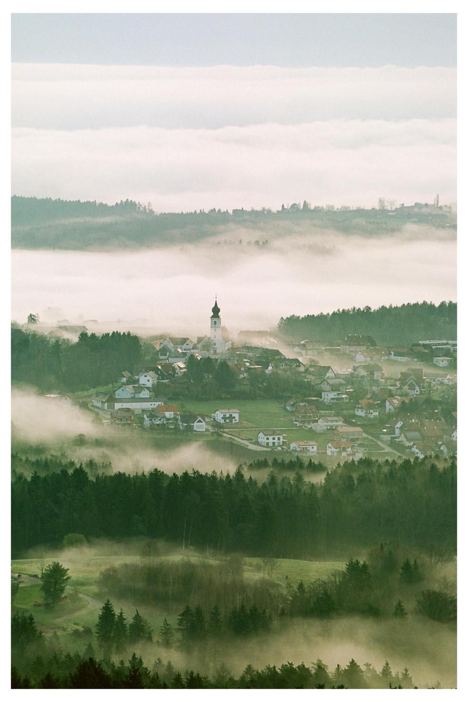 A village with a church steeple is nestled among rolling hills and forests, partially obscured by a layer of fog.