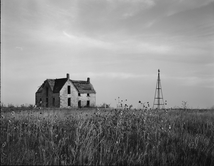 An abandoned stone house and a windmill stand in a field of tall grass and wildflowers under a cloudy sky.