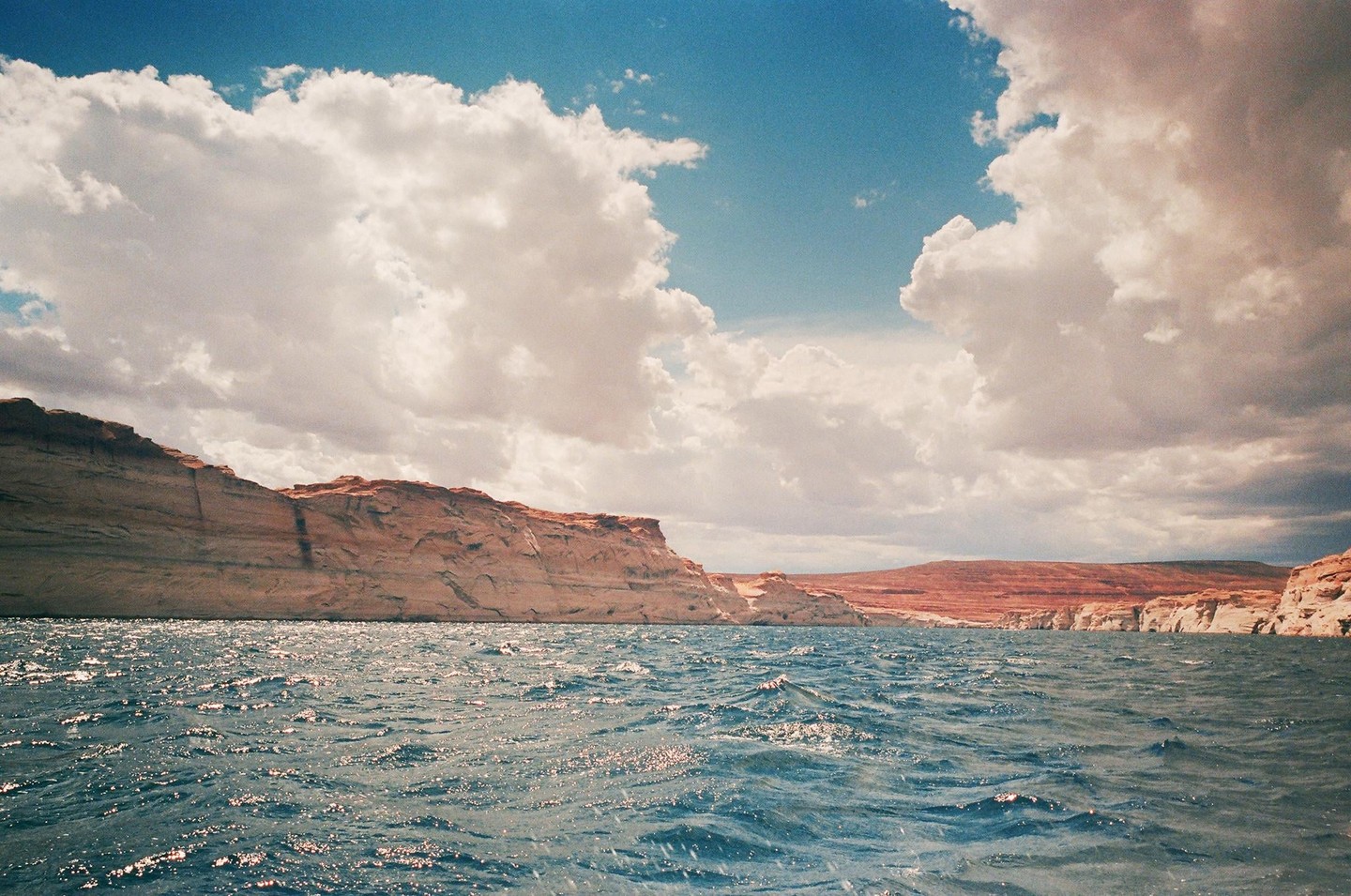 A wide body of water with choppy waves reflects the light from a cloudy sky, with rocky cliffs and hills in the background.