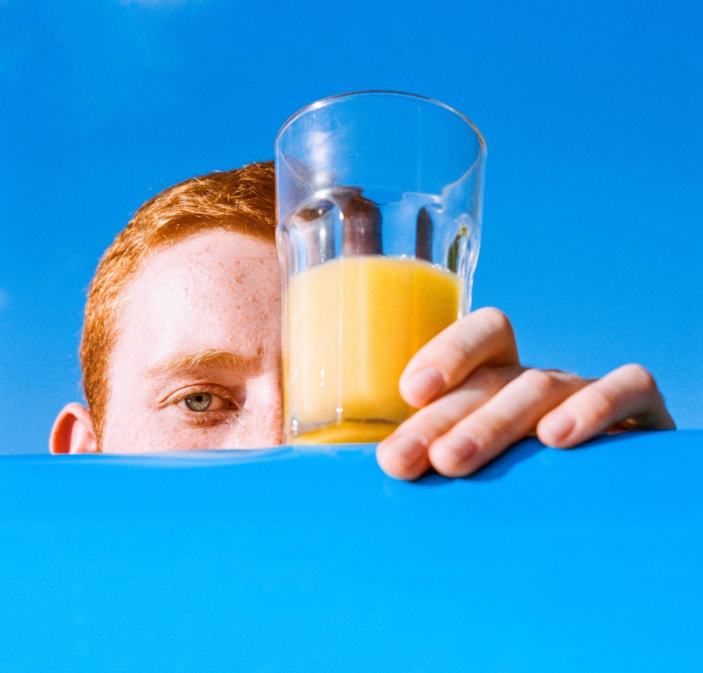 A man with red hair holds a glass of orange juice over a blue surface, with a blue sky in the background.